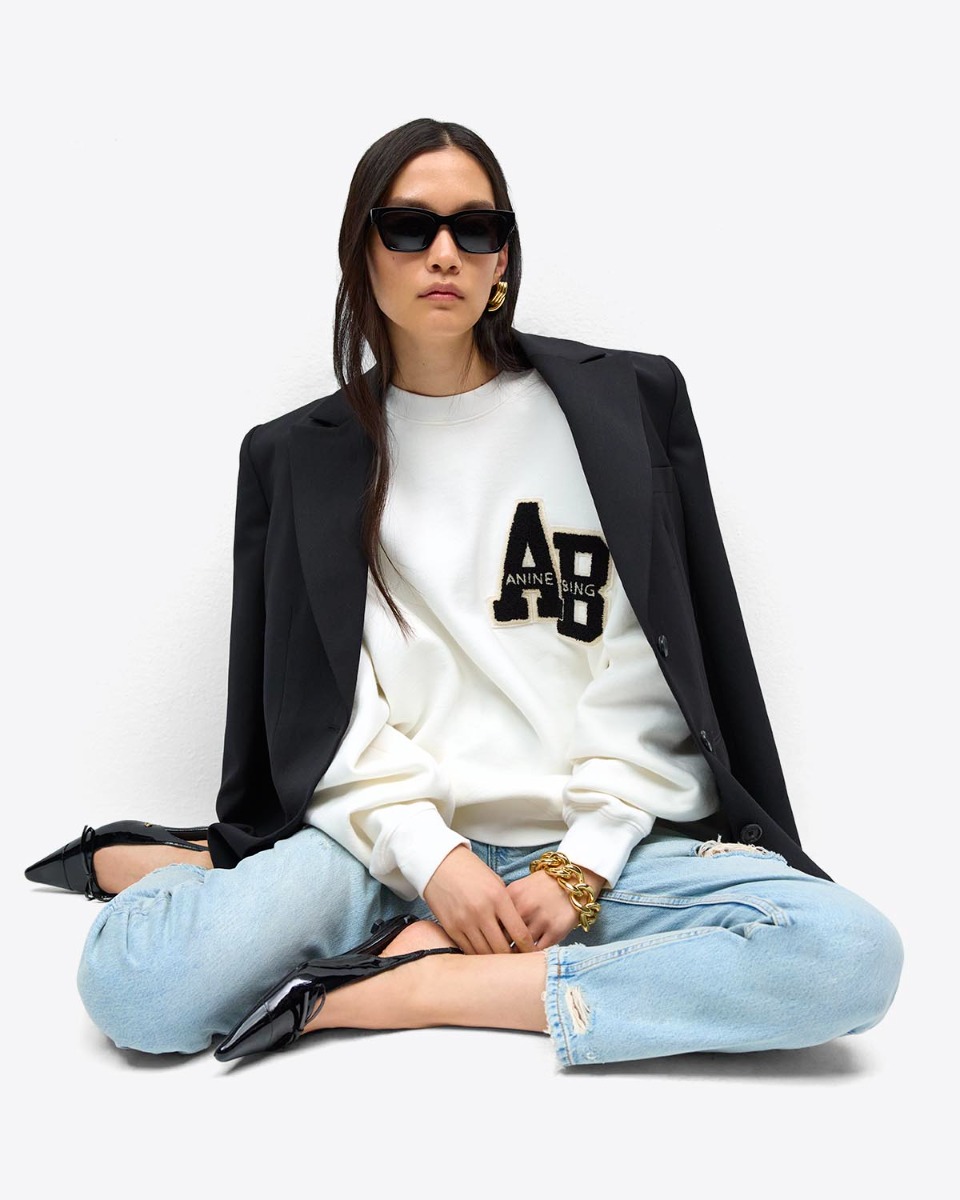 Sweatshirt oversize Miles Letterman Anine Bing en coton blanc logo &laquo; AB &raquo; en chenille noir et &eacute;cru sur la poitrine, port&eacute; avec un jean bleu clair.