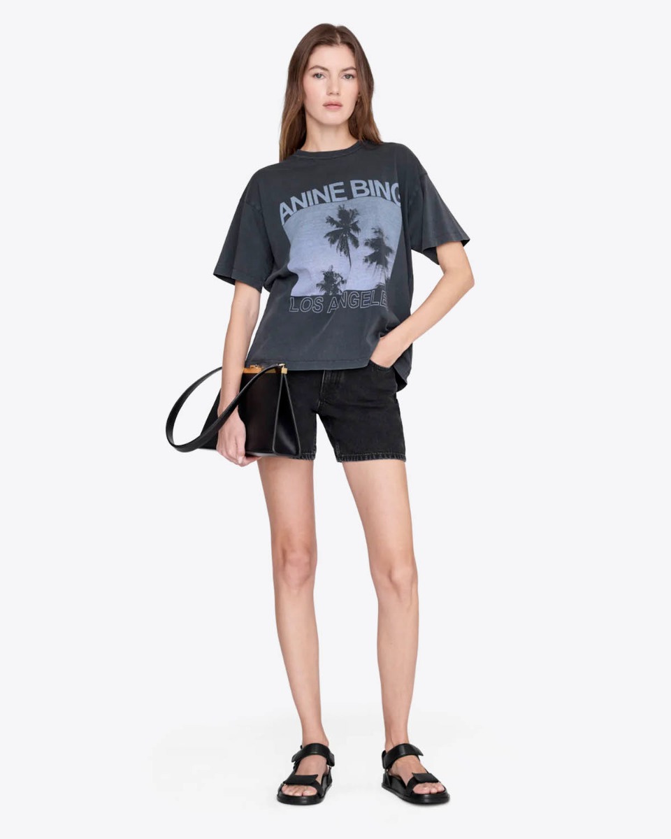 T-shirt manches courtes Cade Palms Anine Bing en jersey de coton fluide noir d&eacute;lav&eacute; s&eacute;rigraphie avec les palmiers mythique de Los Angeles fond bleu et logo ANINE BING, port&eacute; de face avec un short en jean noir.