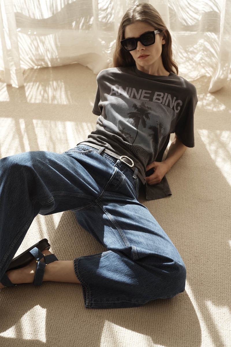 T-shirt manches courtes Cade Palms Anine Bing en jersey de coton fluide noir d&eacute;lav&eacute; s&eacute;rigraphie avec les palmiers mythique de Los Angeles fond bleu et logo &laquo; ANINE BING &raquo;, port&eacute; de face avec un jean bleu.