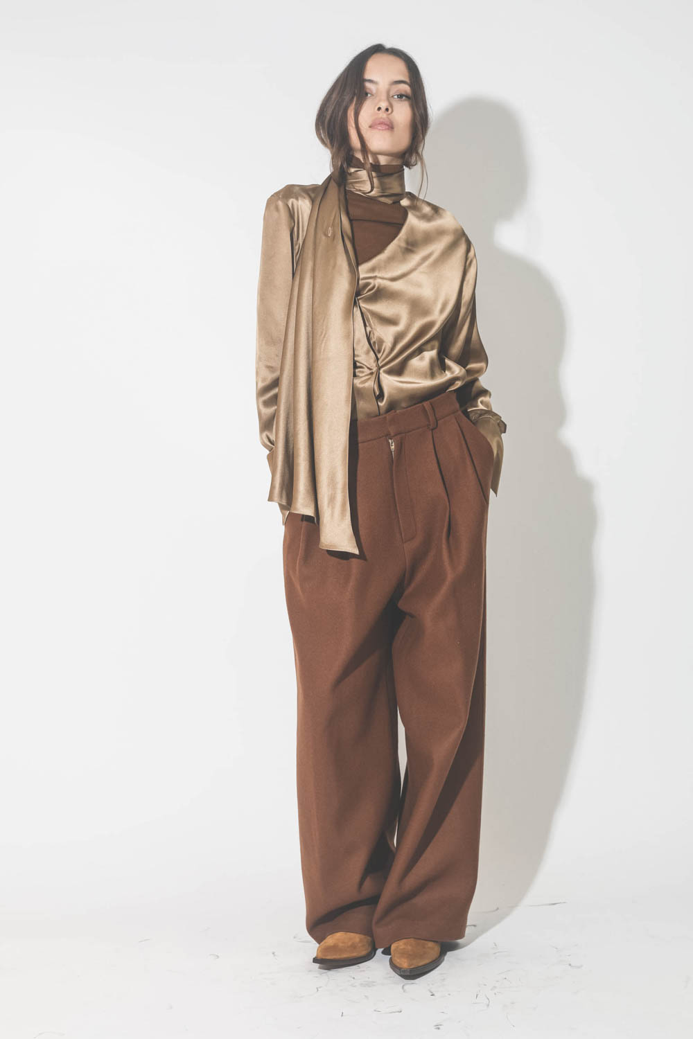Chemise manches longues drapée col écharpe en satin de soie camel doré. Porté avec un pantalon à pinces en drap de laine marron.