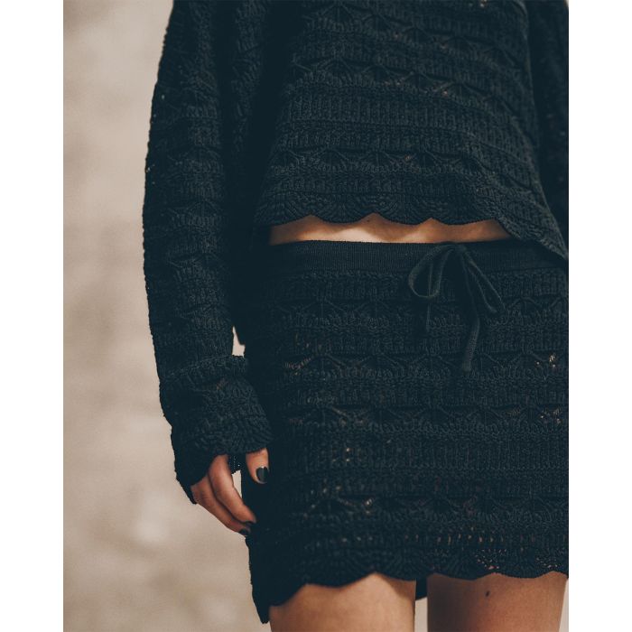 Mini-jupe taille basse en crochet noir en lin et coton Mikuta. Porté de face avec un débardeur en crochet noir assorti. Détail du lien de serrage à la taille.