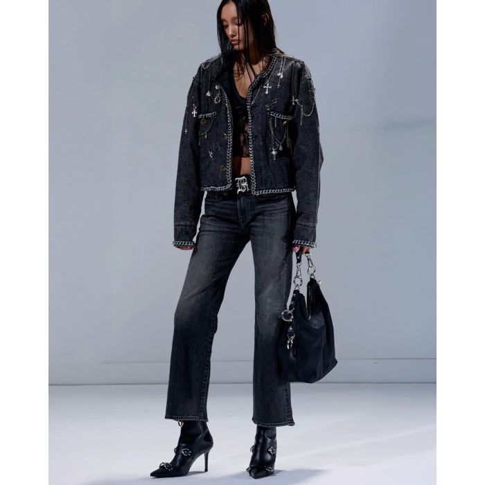 Veste courte Chain Embellished Cropped Chore Jacket R13 en denim gris foncé, customisée avec des chaînes, des épingles à nourrice et des médailles en métal argenté. Portée de face avec un jean noir délavé et des boots à talons.