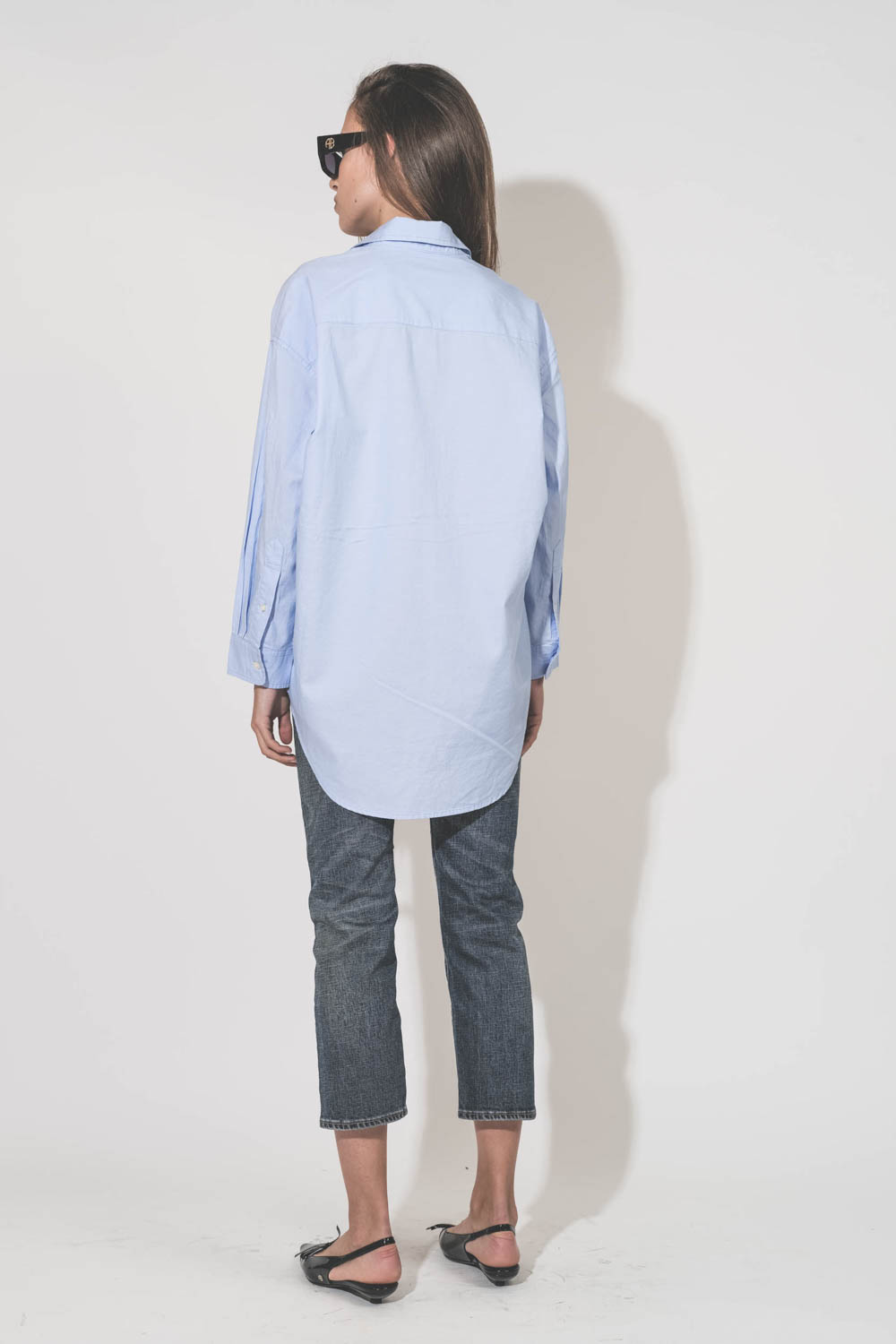 Chemise oxford classique plus longue derrière que devant Button Front bleu ciel Denimist. Porté de dos avec un jean Boy Straight Mire Blue Stretch R13 et des ballerines en cuir noir vernis Slingback Hells Zoey Anine Bing. Lunettes de soleil montures noire