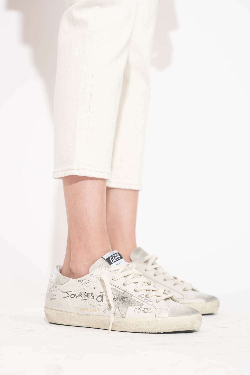 Sneakers en cuir blanc Graffiti écritures et dessins noirs arrière en cuir argent Golden Goose femme. Edition limitée. Portées de profil avec un jean Boy Straight écru stretch R13.