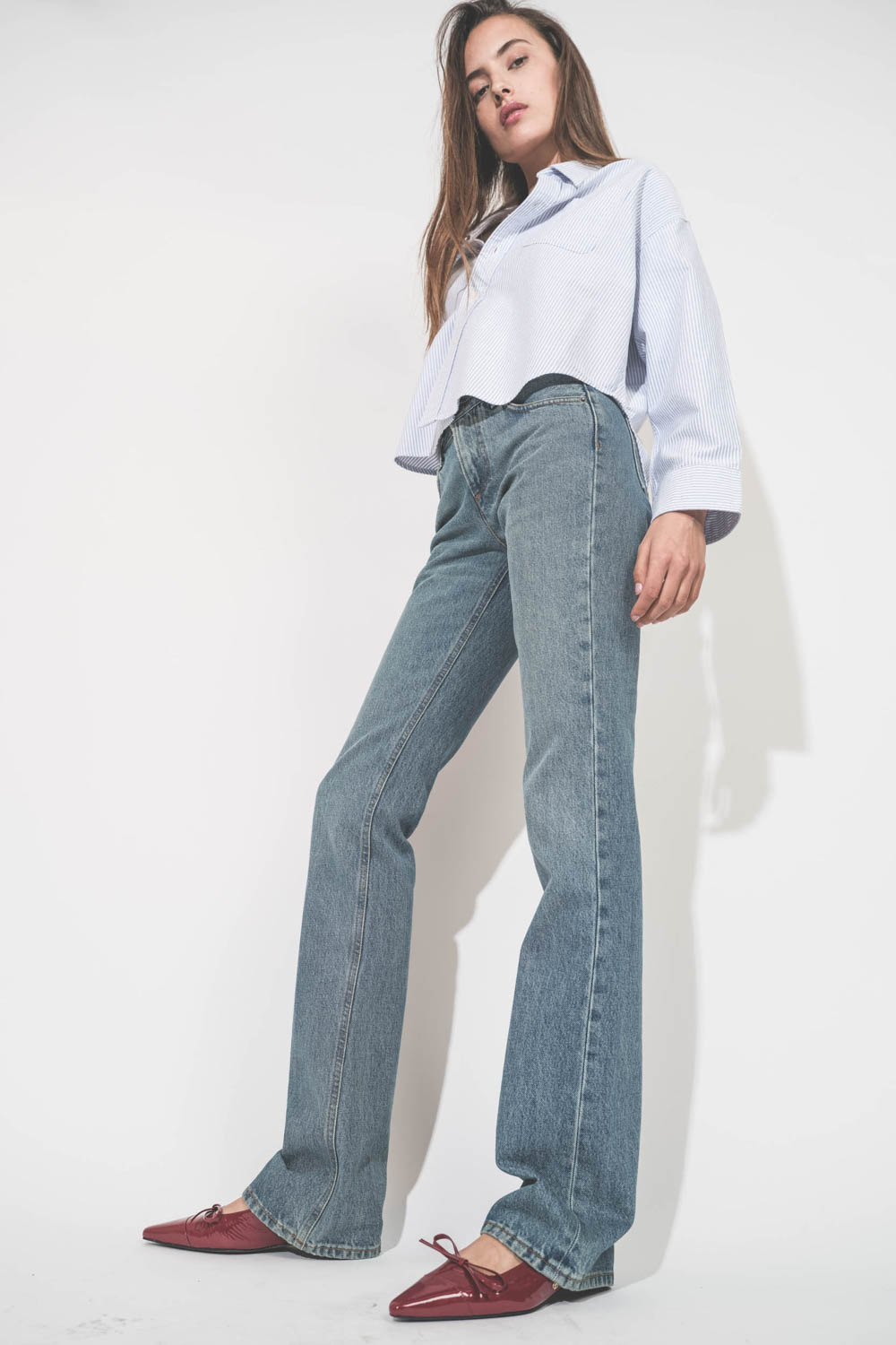 Jean droit taille basse et long bleu délavé Carly Haikure. Porté de profil avec une chemise courte à rayures bleues Button Front Denimist et des ballerines rouge Zoey Anine Bing. Collier Adriel rouge Isabel Marant.
