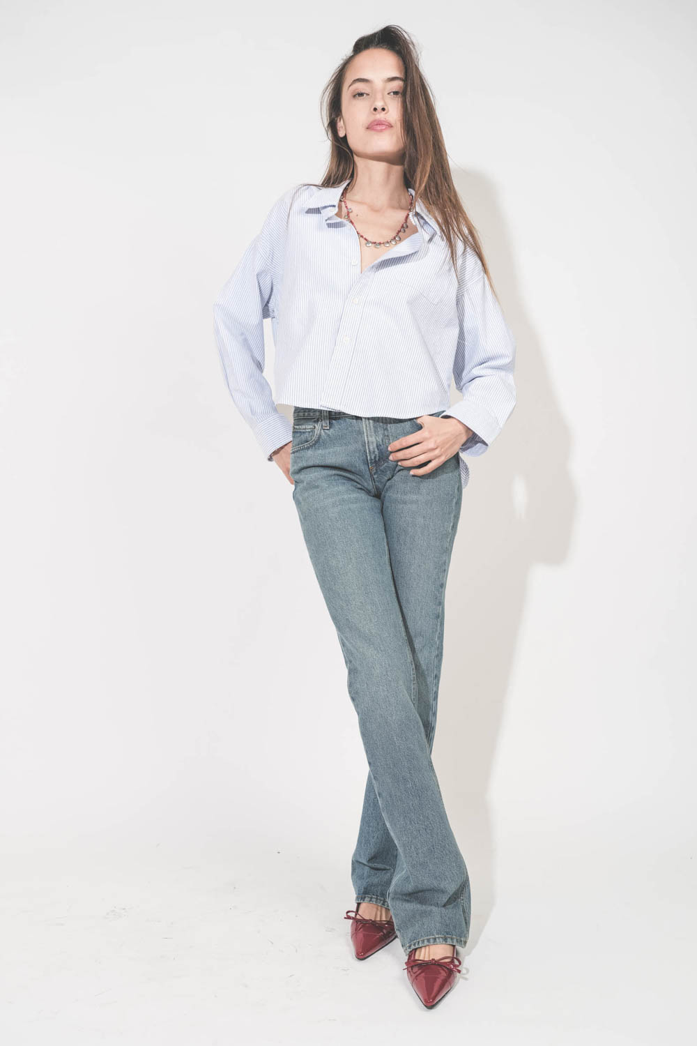 Jean droit taille basse et long bleu délavé Carly Haikure. Porté de face avec une chemise courte à rayures bleues Button Front Denimist et des ballerines rouge Zoey Anine Bing. Collier Adriel rouge Isabel Marant.