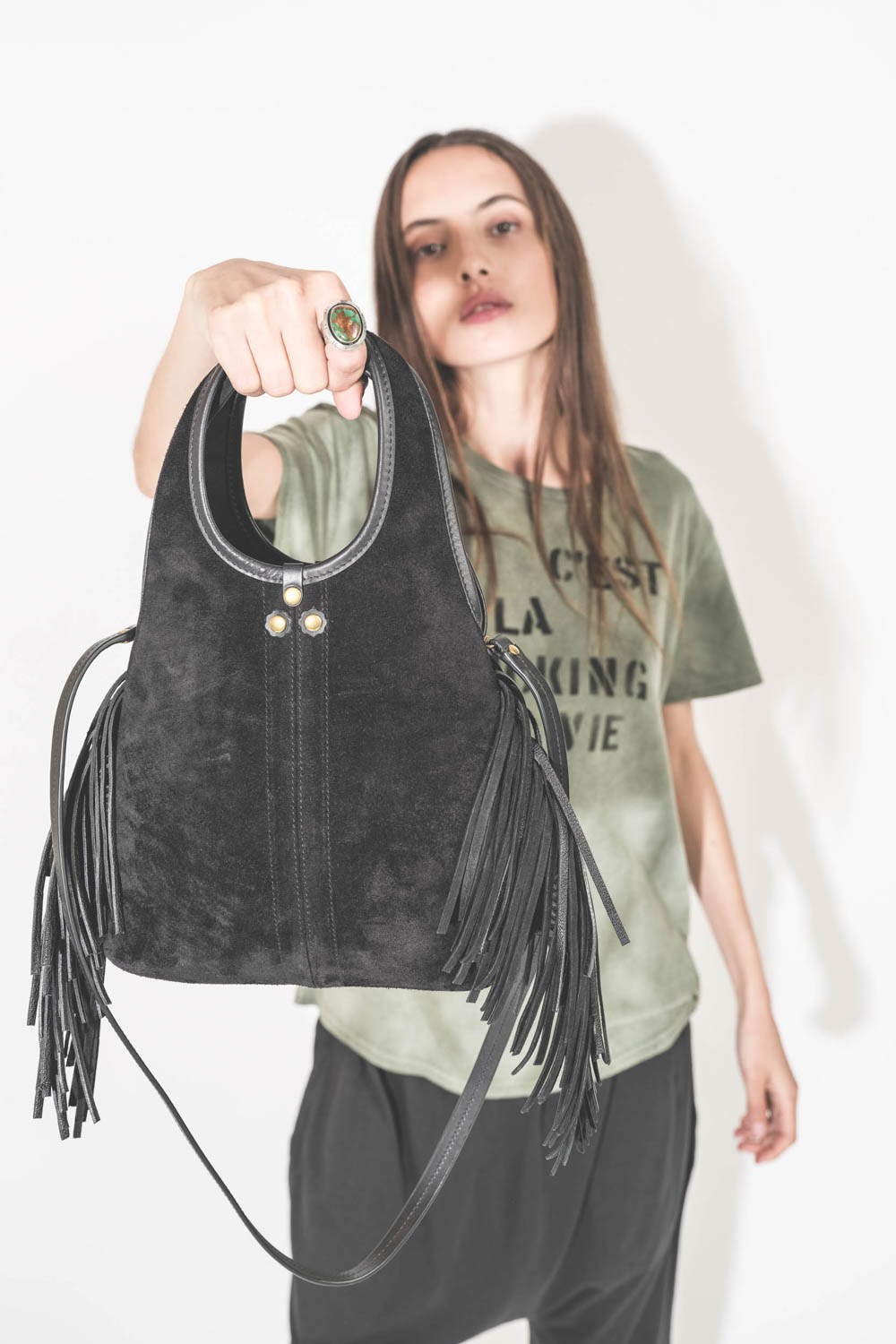 Mini-sac bandoulière en cuir suédé à franges Paco S Jérôme Dreyfuss. Collection capsule SS25. Porté avec un tee-shirt kaki "C'est la Fucking Vie" R13 et un pantalon sarouel en jersey noir The Great. Bague argent et turquoise Harpo. 