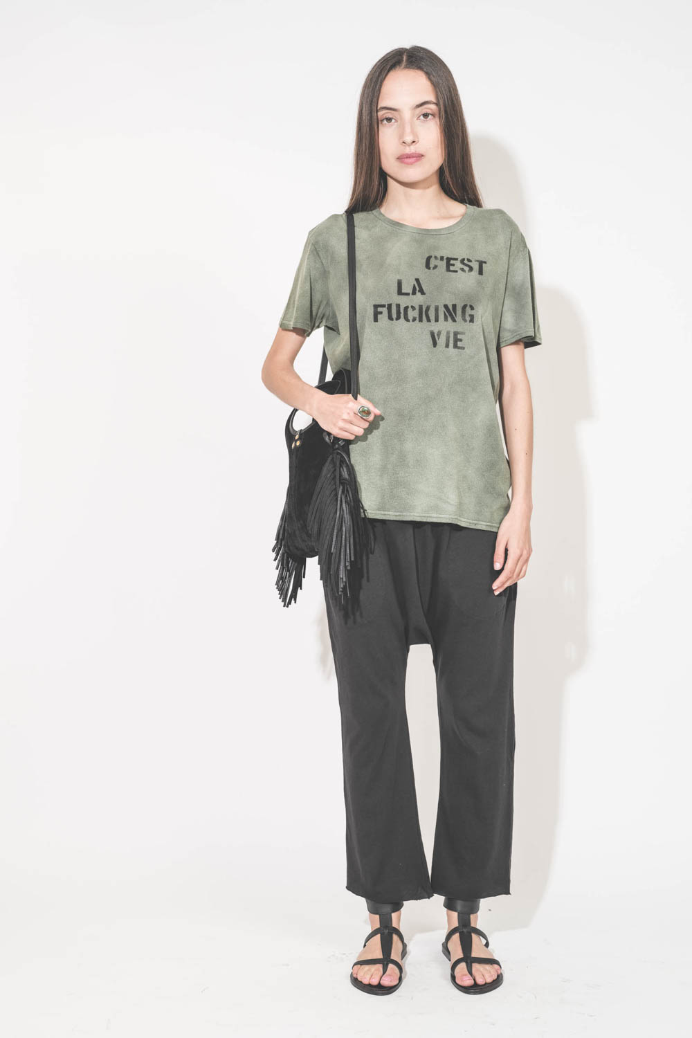 Mini-sac bandoulière en cuir suédé à franges Paco S Jérôme Dreyfuss. Collection capsule SS25. Porté avec un tee-shirt kaki "C'est la Fucking Vie" R13 et un pantalon sarouel en jersey noir The Great. Sandales en cuir noir Ebene semelles noires Caravelle K.
