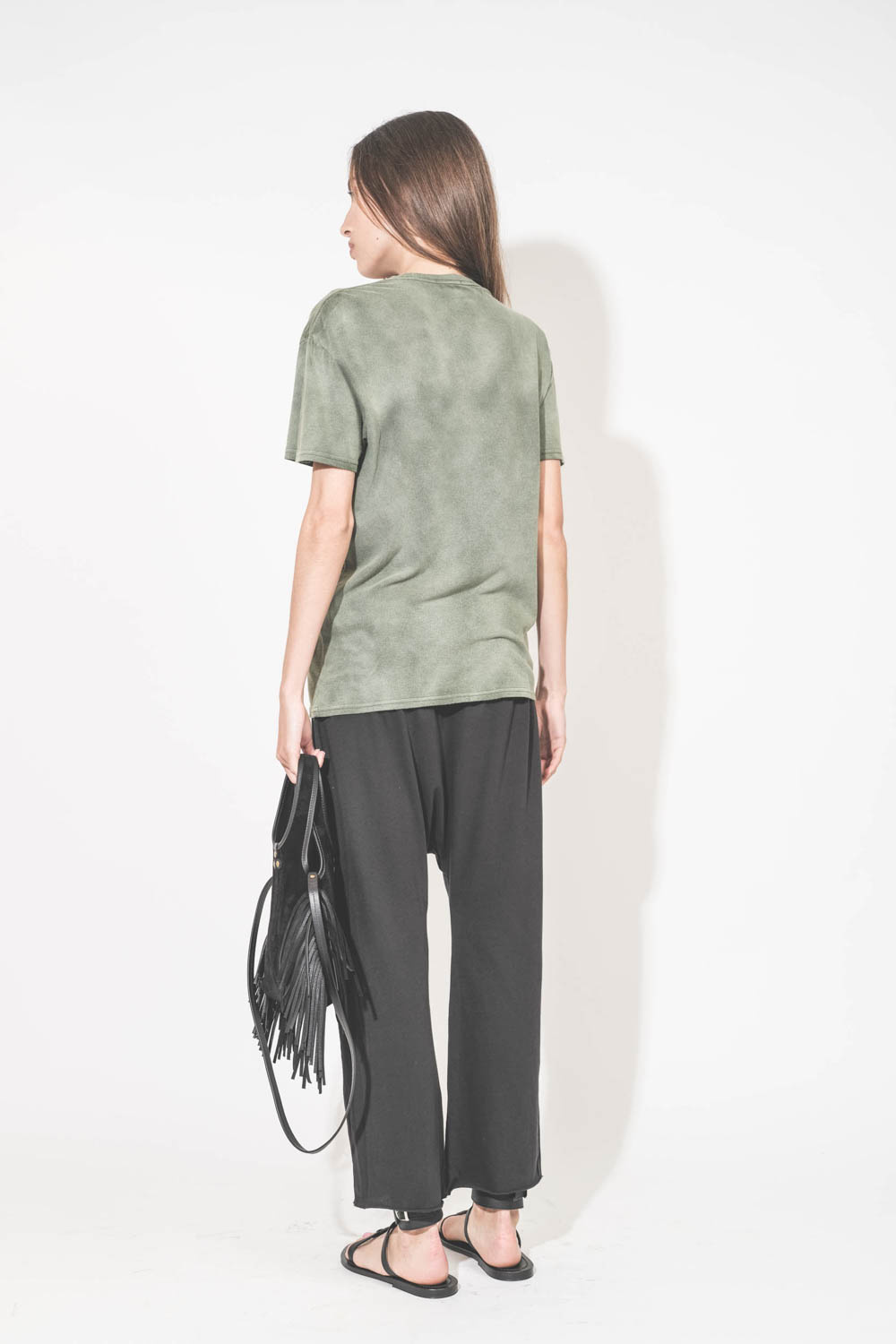 Mini-sac bandoulière en cuir suédé à franges Paco S Jérôme Dreyfuss. Collection capsule SS25. Porté de dos avec un tee-shirt kaki "C'est la Fucking Vie" R13 et un pantalon sarouel en jersey noir The Great. Sandales en cuir noir Ebene semelles noires Carav