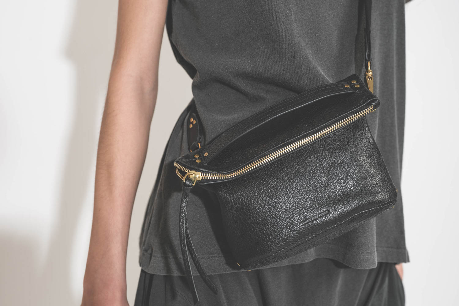 Pochette en cuir de chèvre noir grainé portée crossbody avec une bandoulière sangle large noire Jérôme Dreyfuss. Tee-shirt manches courtes Boxy Crew et pantalon sarouel en jersey noir délavé The Great. Détail de la pochette. 