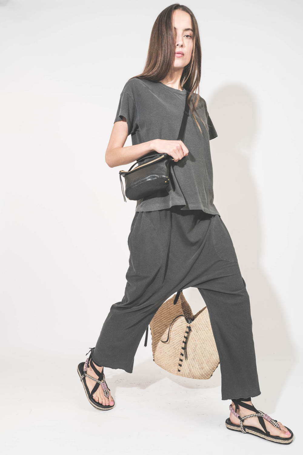 Pochette en cuir de chèvre noir grainé portée de profil crossbody avec une bandoulière sangle large noire Jérôme Dreyfuss. Tee-shirt manches courtes Boxy Crew et pantalon sarouel en jersey noir délavé The Great. Sandales en cordes tressées et ruban noir G