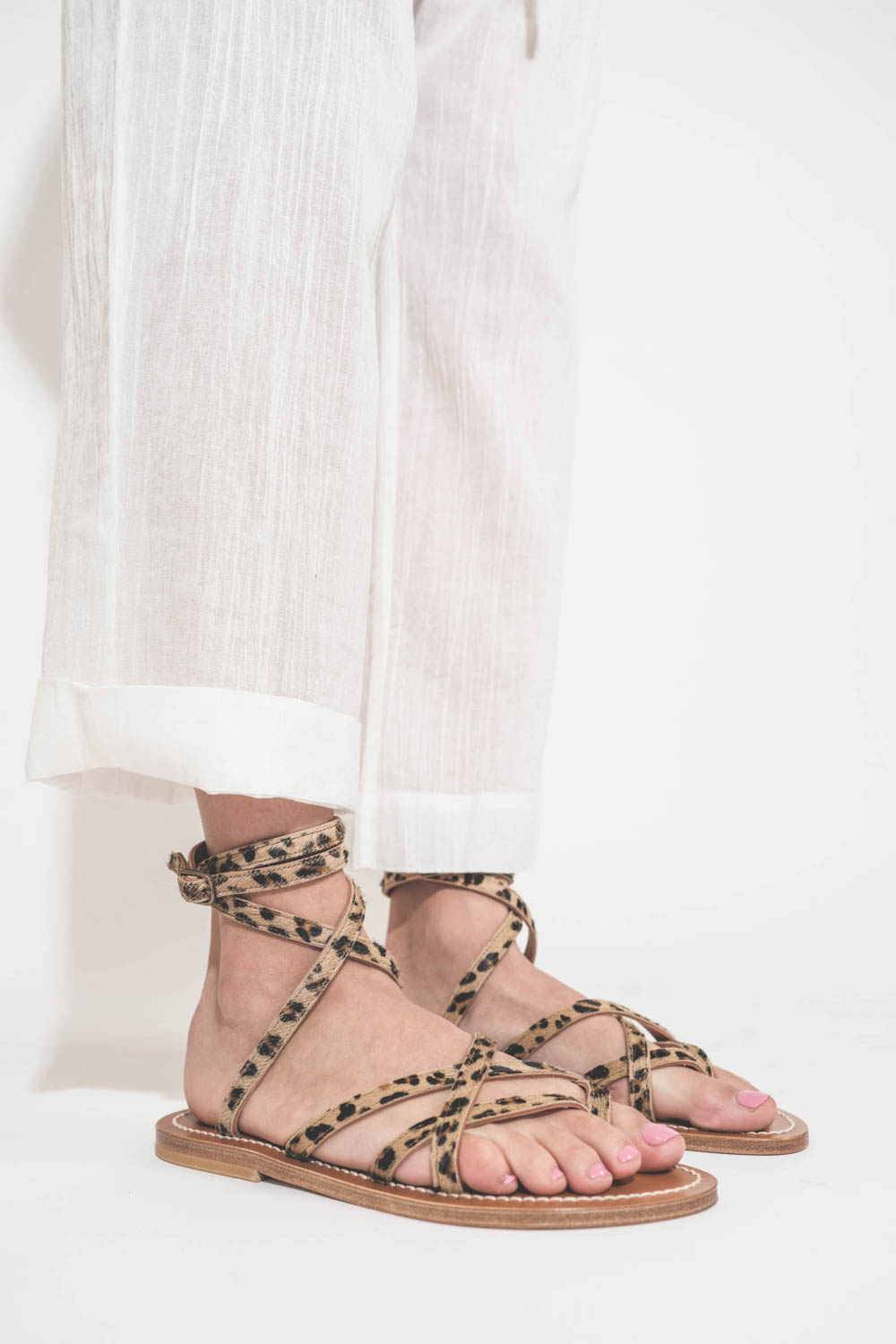 Sandales plates avec brides Zenobie K.Jacques St-Tropez en cuir "pony" léopard. Porté de profil avec un pantalon en voile de coton blanc The Great.