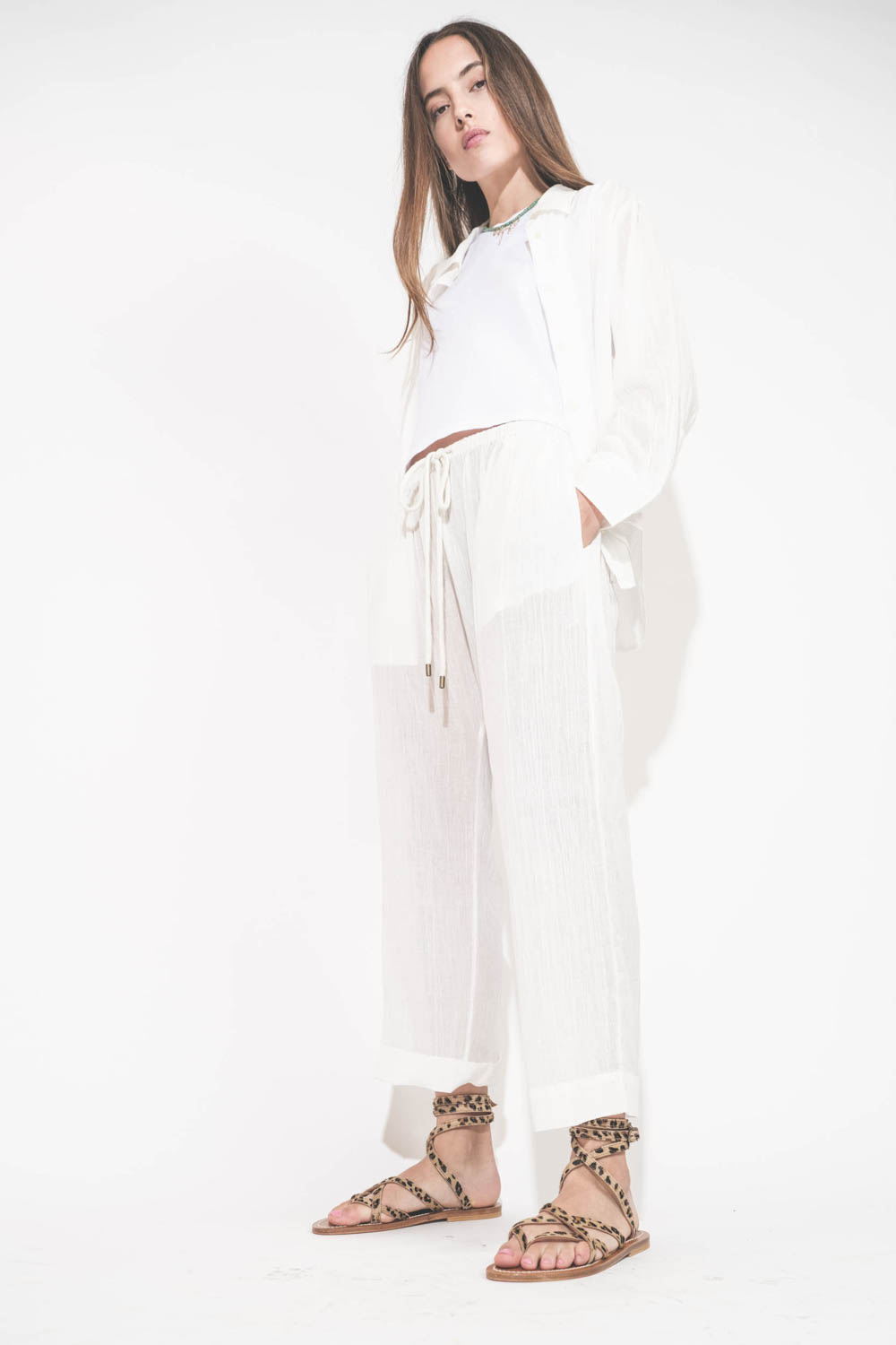 Sandales plates avec brides Zenobie K.Jacques St-Tropez en cuir "pony" léopard. Porté de profil avec un chemise en voile de coton blanc et un pantalon assorti en voile de coton blanc The Great. 