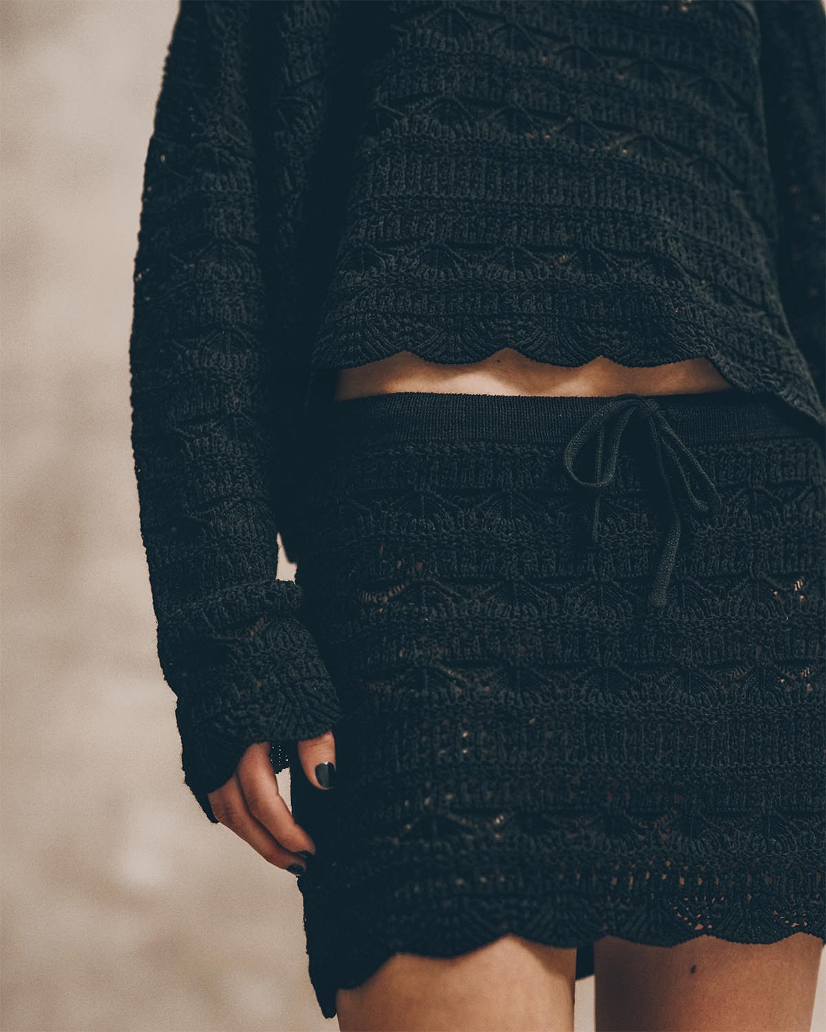 Mini-jupe taille basse en crochet noir en lin et coton Mikuta. Porté de face avec un débardeur en crochet noir assorti. Détail du lien de serrage à la taille.