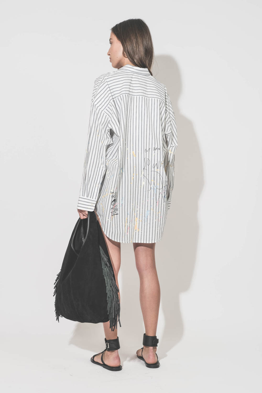 Chemise oversized en popeline blanche à rayures noires et graffiti Drop Neck R13. Portée de dos avec des sandales noires Caravelle K.Jacques St Tropez et un sac à franges Paco Jérôme Dreyfuss.
