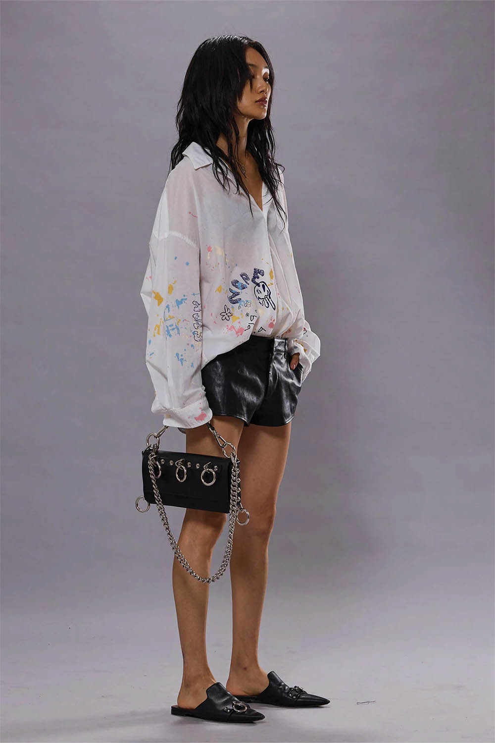 Chemise manches longues oversize en coton blanc avec dessins esprit graffiti all over sur tout le bas de la chemise Drop Neck shirt R13. Porté avec un mini-short en cuir noir et des mules. 