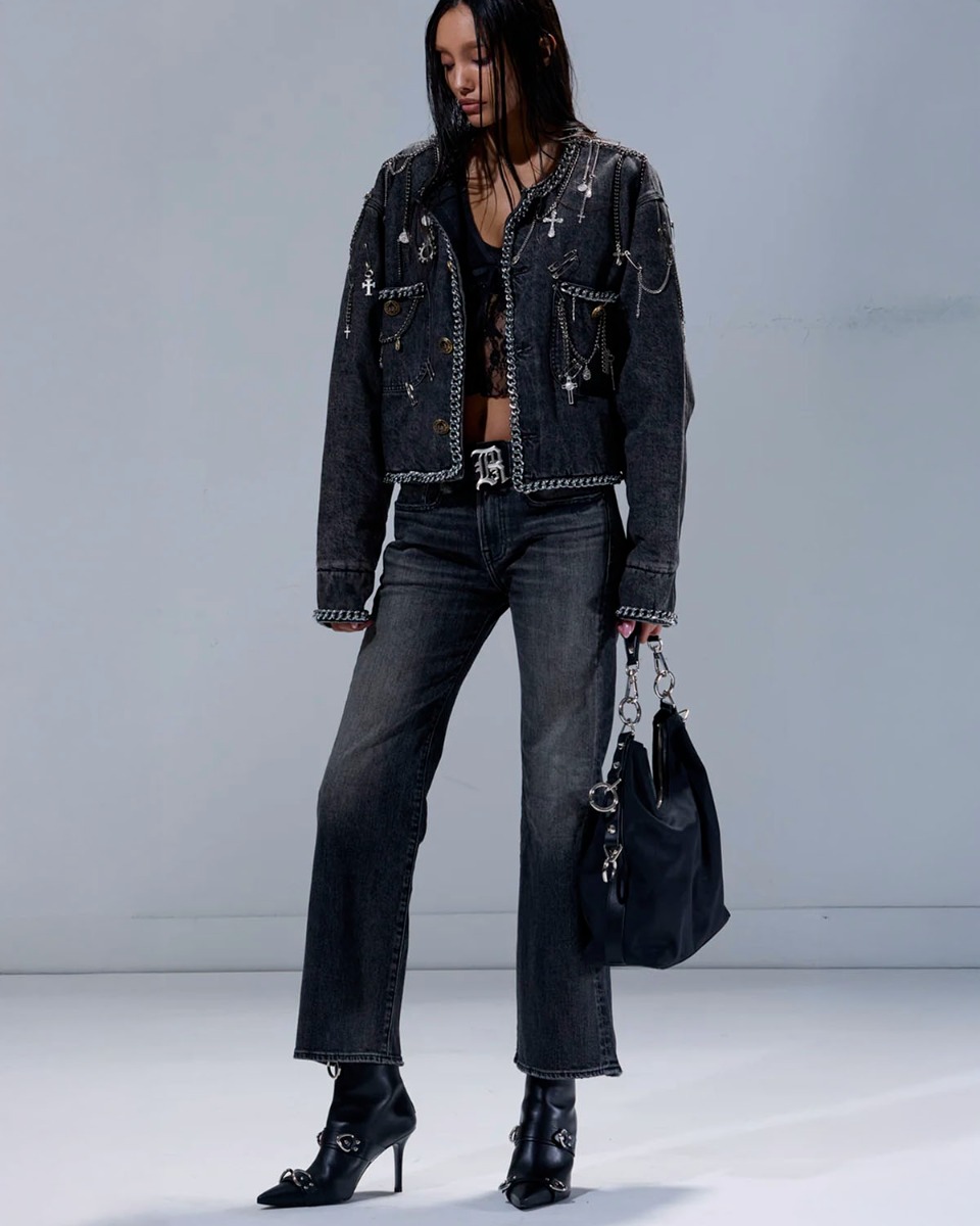 Veste courte Chain Embellished Cropped Chore Jacket R13 en denim gris foncé, customisée avec des chaînes, des épingles à nourrice et des médailles en métal argenté. Portée de face avec un jean noir délavé et des boots à talons.