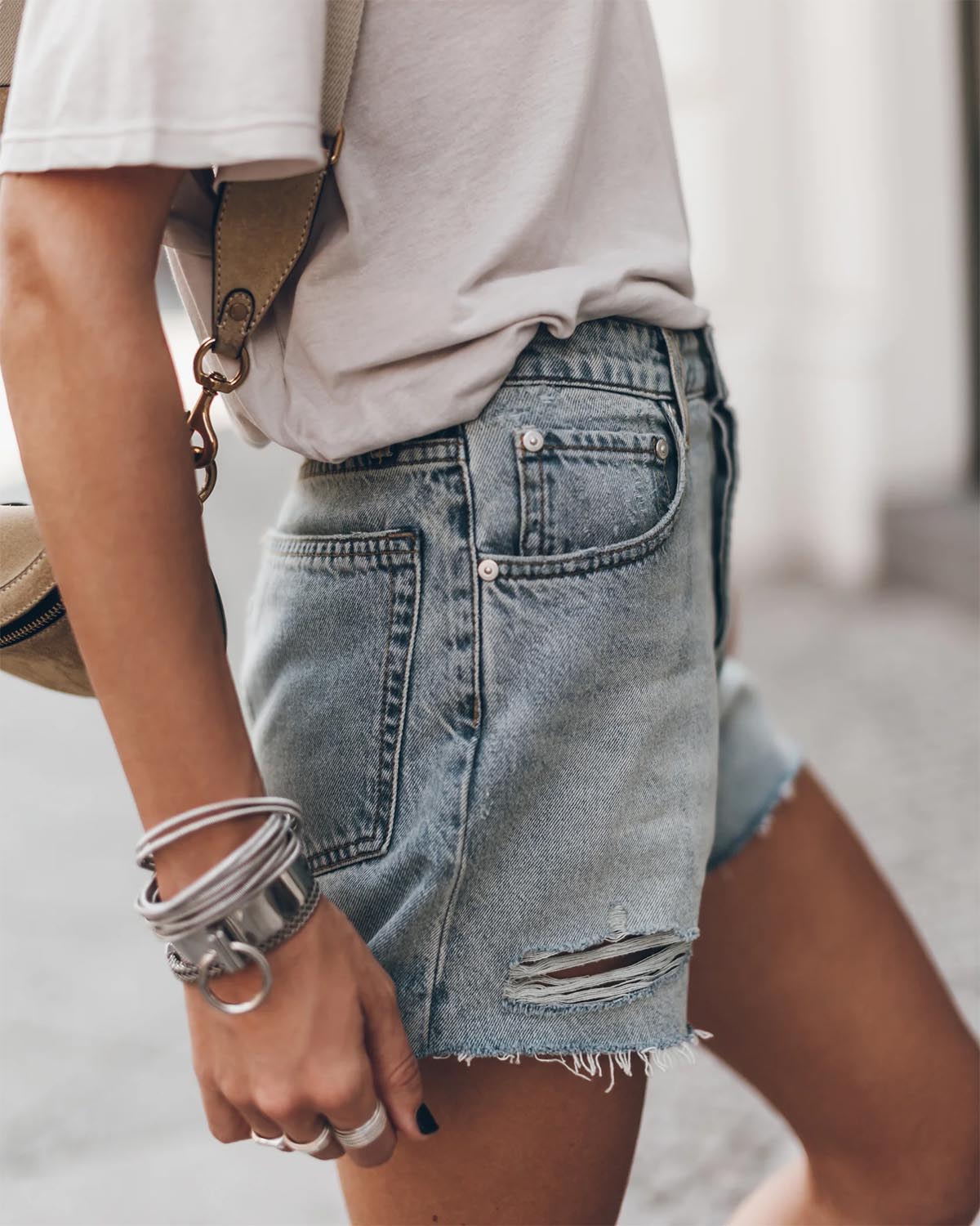 Short en jean bleu ciel usé Mikuta. Porté de profil avec un tee-shirt écru Mikuta. Bracelets en argent Mikuta.