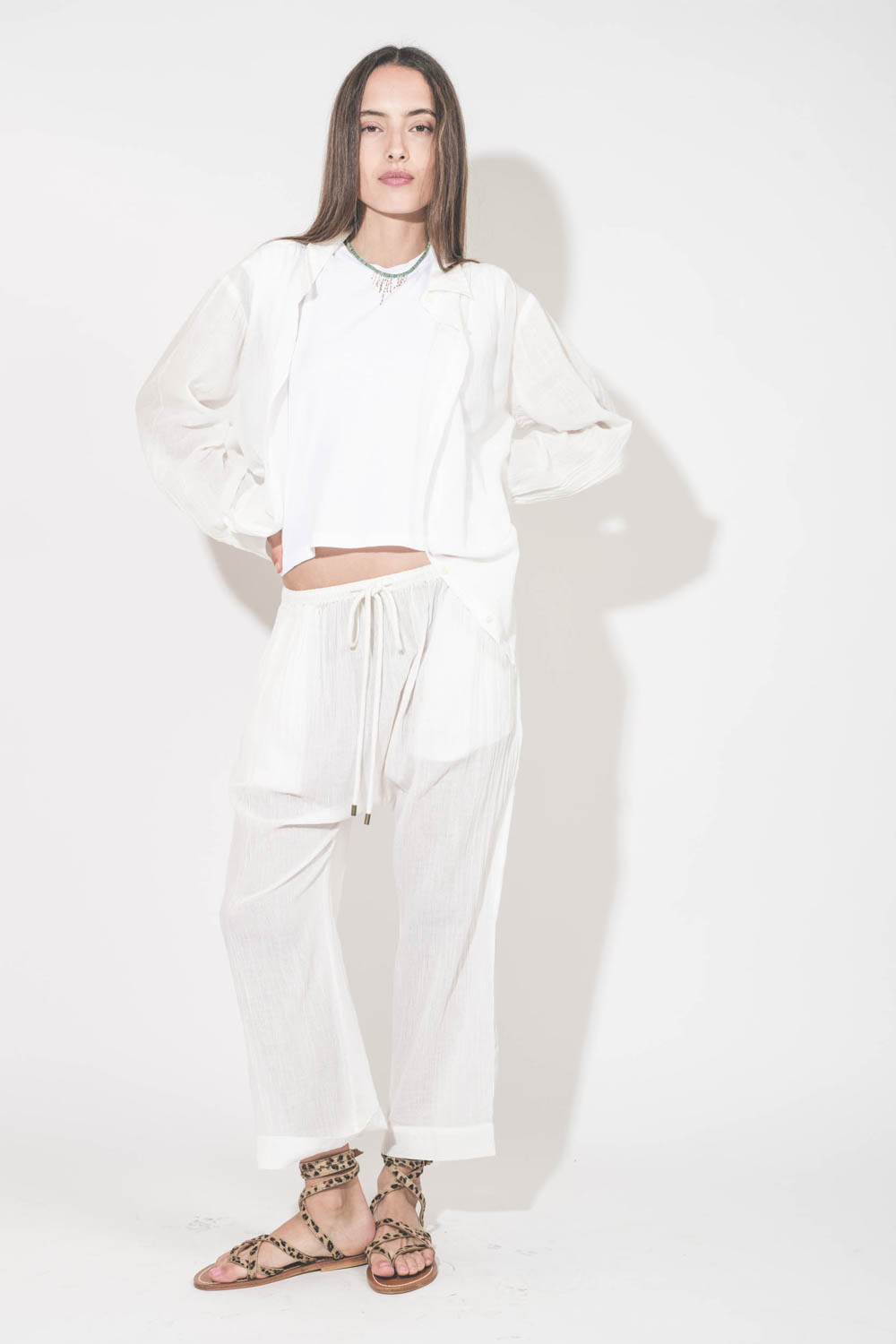 Pantalon Sarouel en voile de coton blanc Reef The Great. Porté de face avec une chemise manches longues en voile de coton blanc et un tee-shirt sans manches Peak Shoulder The Great. Sandales en cuir pony léopard Zenobie K.Jacques St-Tropez. Collier turquo