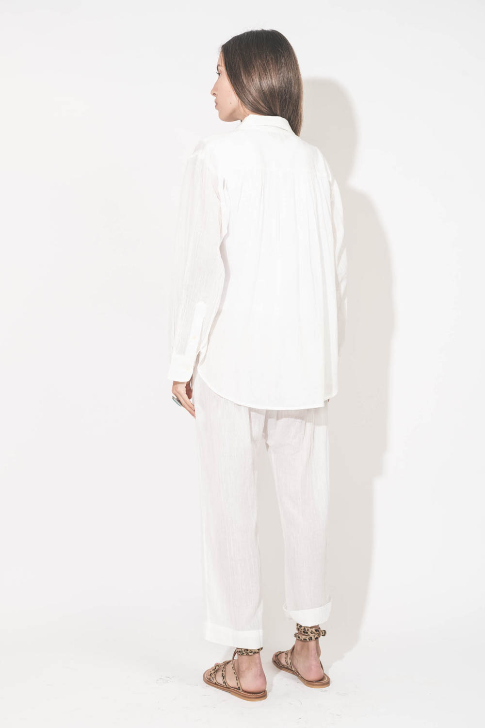 Pantalon Sarouel en voile de coton blanc Reef The Great. Porté de dos avec une chemise manches longues en voile de coton blanc et un tee-shirt sans manches Peak Shoulder The Great. Sandales en cuir pony léopard Zenobie K.Jacques St-Tropez. Collier turquoi