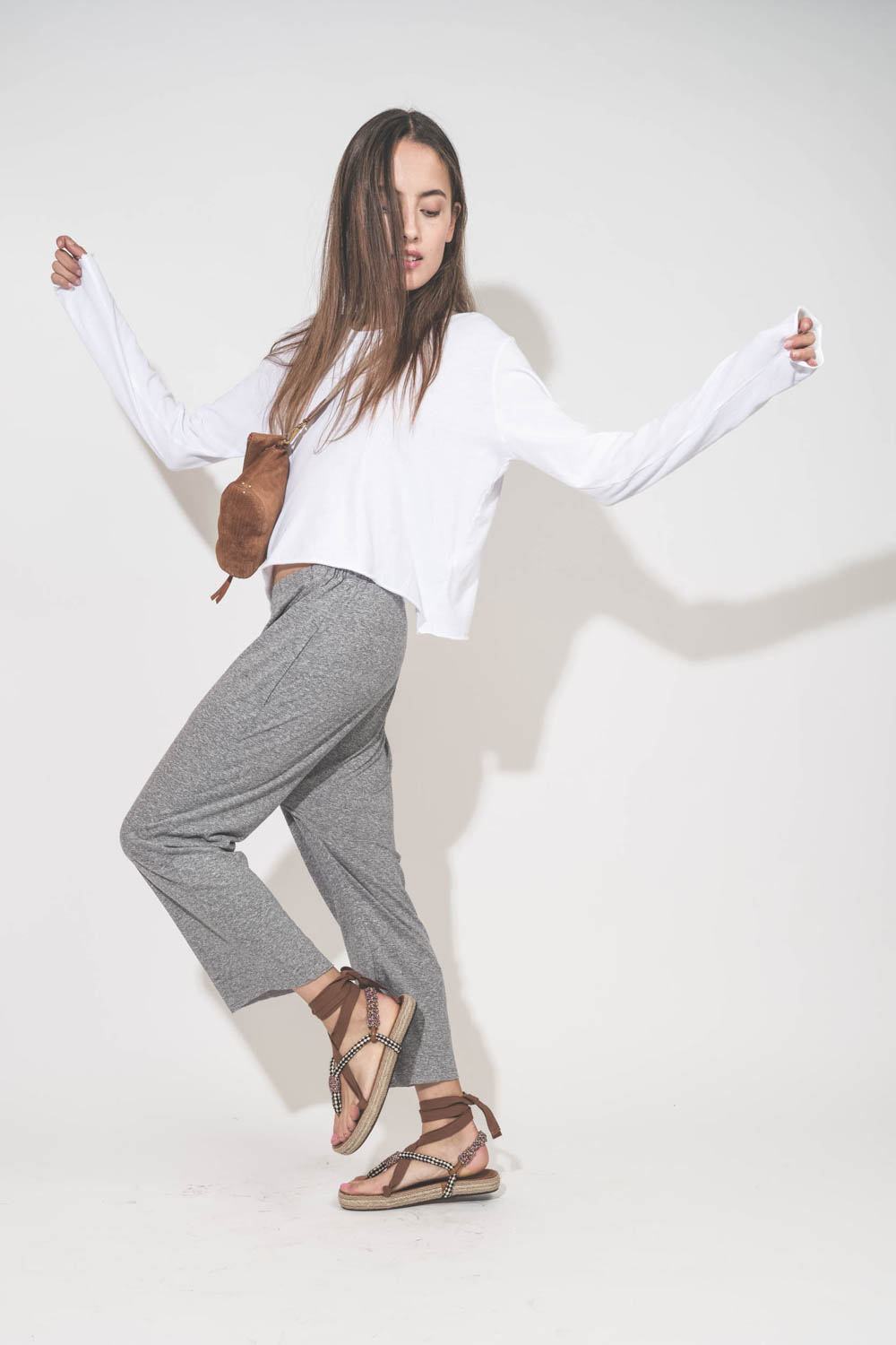 Pantalon sarouel court en jersey gris chiné The Great. Porté de profil avec une tee-shirt court manches longues blanc et des sandales en cordes tressées et ruban beige Gypsa Isabel Marant. Pochette Lucky en cuir suédé caramel portée crossbody avec une san