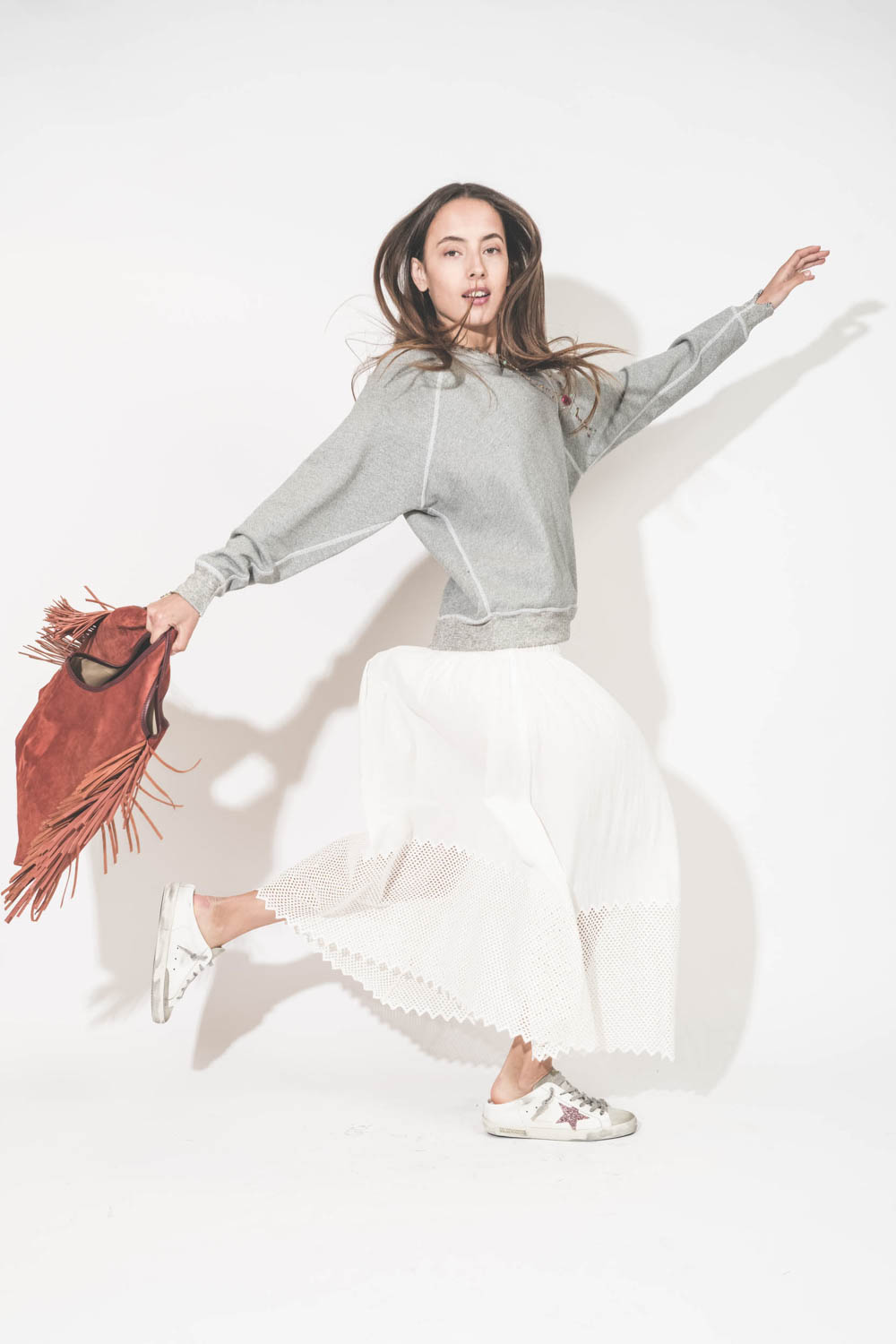 Jupe longue en coton et nid d'abeille placé sur le bas de la jupe blanche The Canary Skirt The Great. Portée de profil avec un sweat-shirt gris chiné College The Great et des sneakers Super Star sabot Golden Goose. Cabas à franges en cuir Terracotta Paco 
