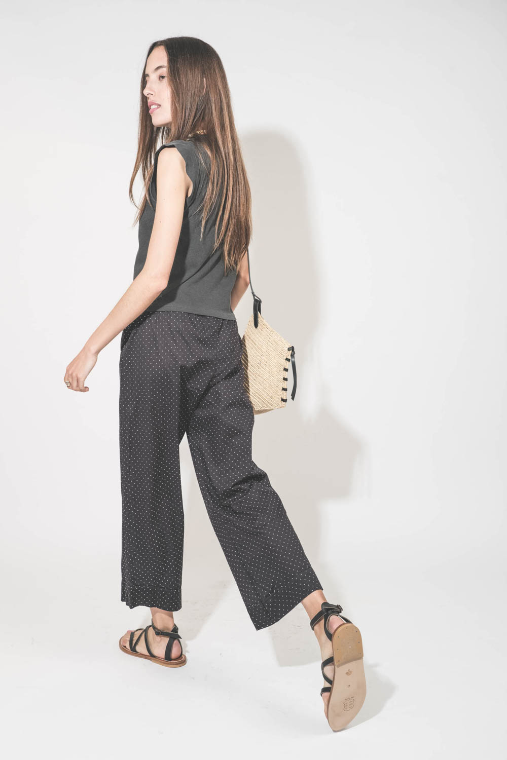 Pantalon taille basse en voile de coton noir à pois blancs Easy Sleep The Great. Porté de profil dos avec un tee-shirt sans manches noir Peak Shoulder The Great et des sandales en cuir noires Zenobie K.Jacques St-Tropez. Panier en osier bandoulière en cui