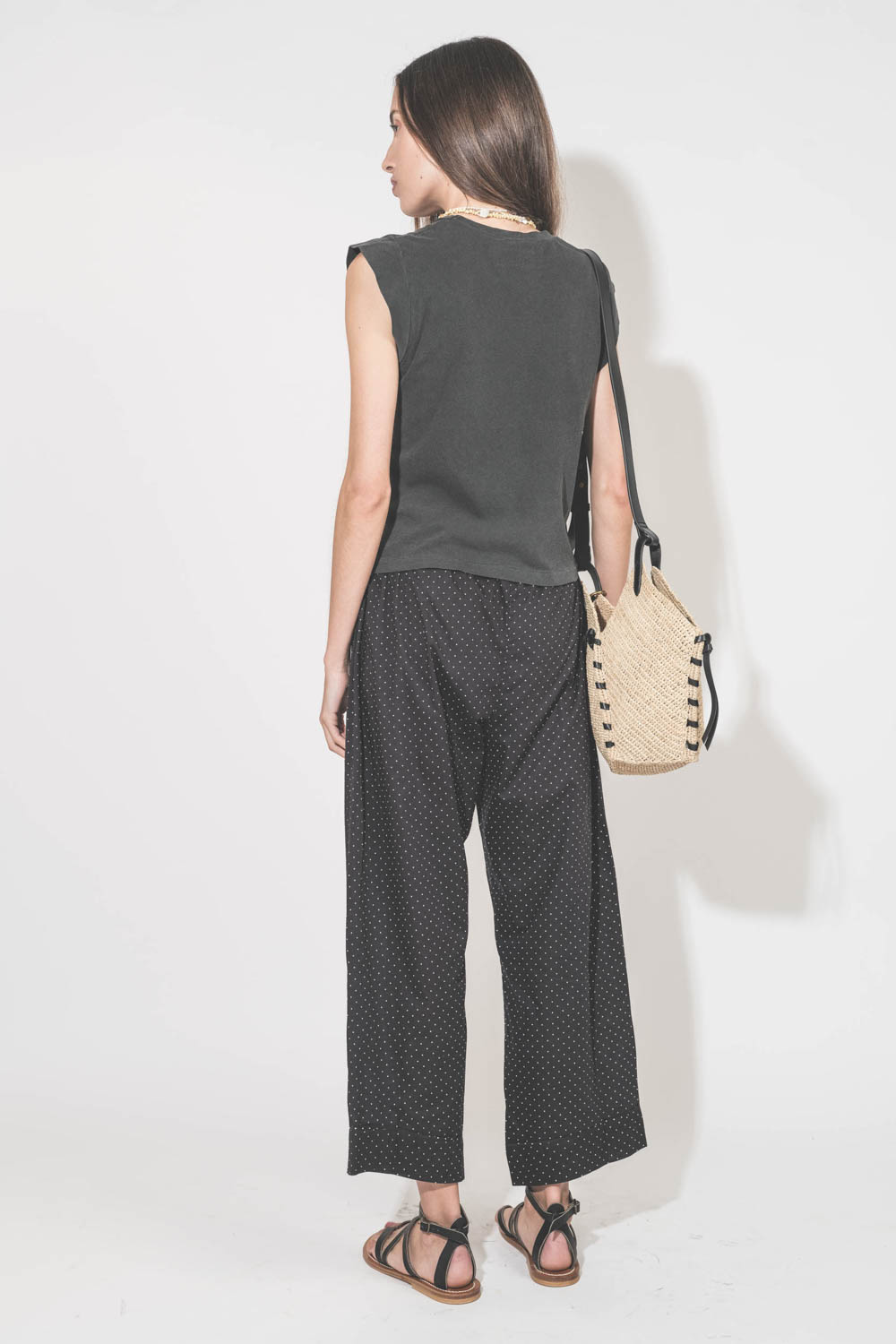 Pantalon taille basse en voile de coton noir à pois blancs Easy Sleep The Great. Porté de dos avec un tee-shirt sans manches noir Peak Shoulder The Great et des sandales en cuir noires Zenobie K.Jacques St-Tropez. Panier en osier bandoulière en cuir noire