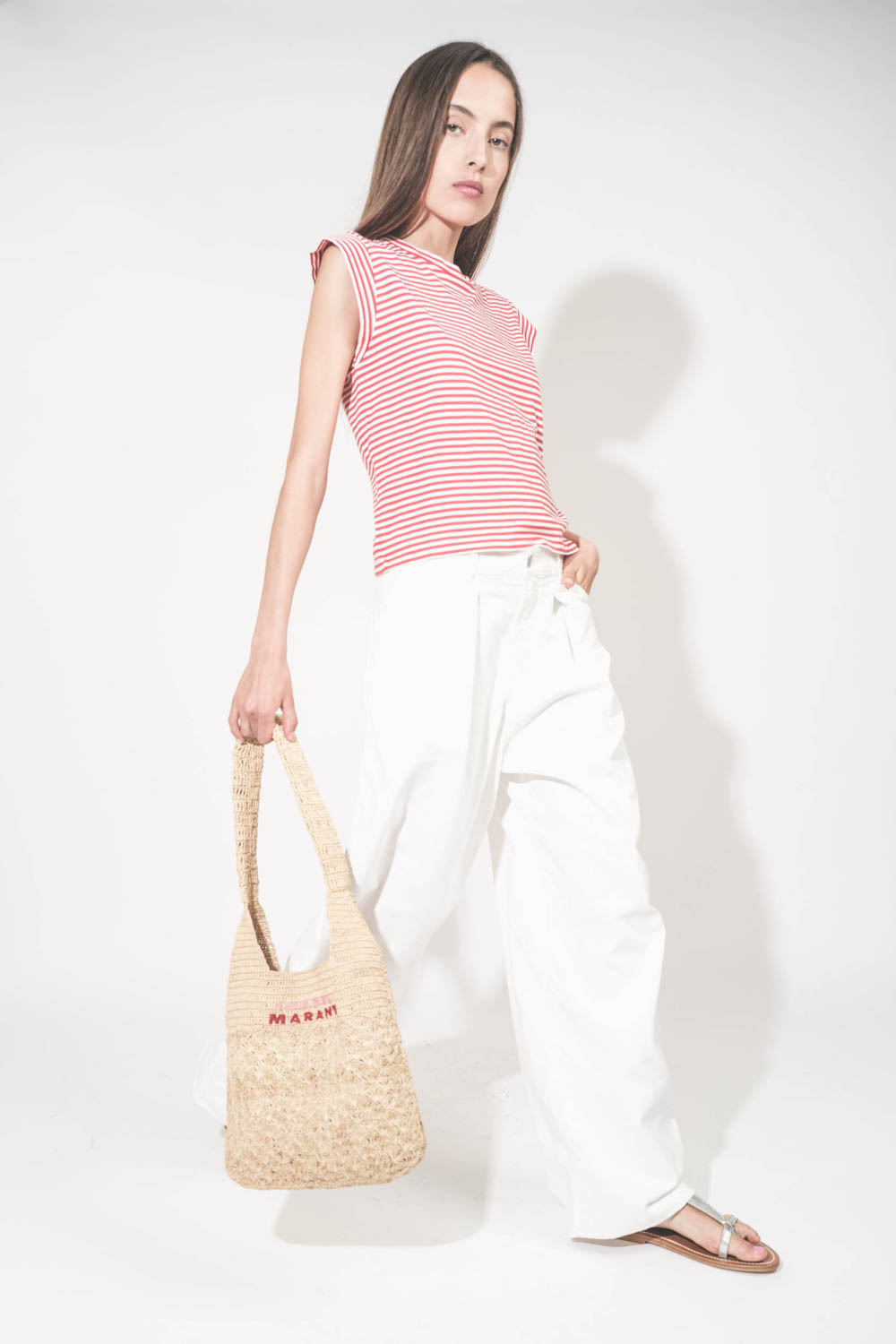 Tee-shirt manches courtes raccourcies en coton rayé rouge et blanc Peak Shoulder The Great. Porté de profil avec un jean large blanc Georgia Denimist et des sandales en cuir argent Caravelle K.Jacques St-Tropez. Panier en osier naturel Praia Isabel Marant
