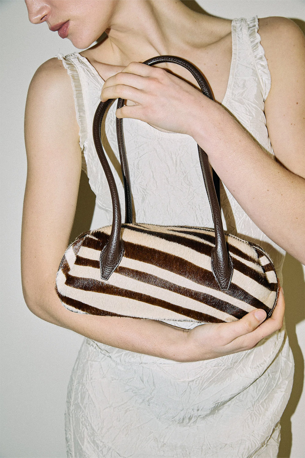 Sac Mini Oyster Yuzefi en cuir pony Savanna imprimé zèbre marron et écru, mini sac baguette porté épaule au design contemporain. Porté dans les bras.